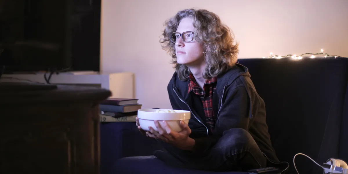 A young person sitting cross-legged in a dim living room at night, holding a bowl of popcorn and watching a screen just out of frame